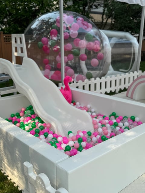 Interactive playground structure with white slide leading to colorful ball pit, pink flowers in dome above, and white picket fence