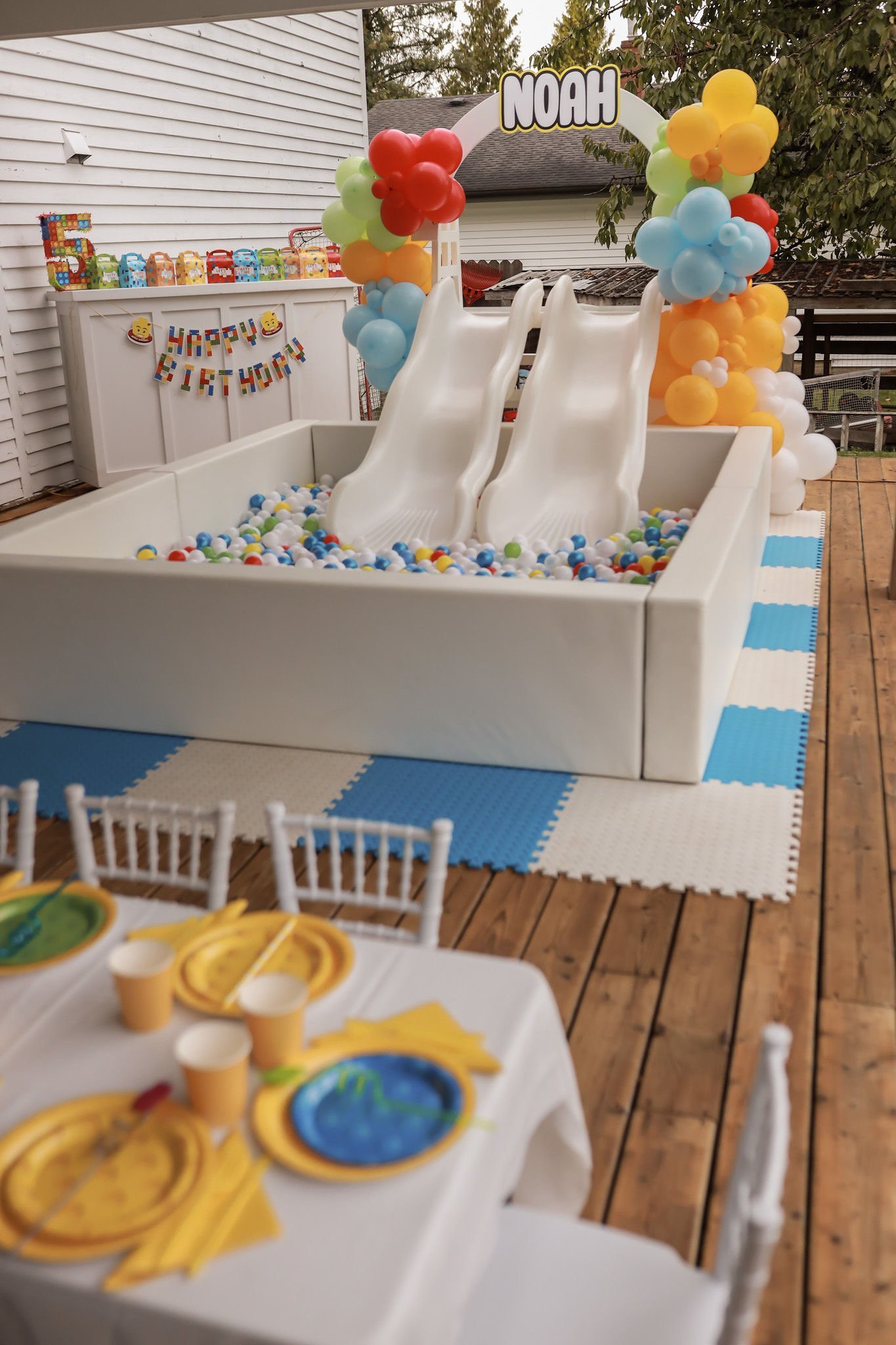 Outdoor birthday party setup with white bounce house filled with colorful balls, balloon arch, Happy Birthday banner, and table setting with yellow plates on wooden deck