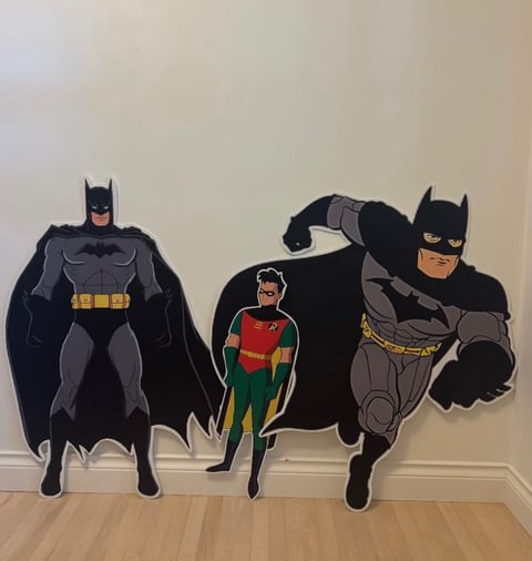 Cardboard cutouts of Batman, Robin, and another Batman figure standing on wooden floor against white wall