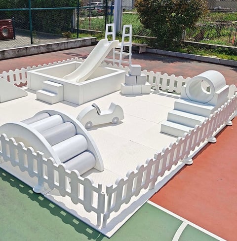 White inflatable playground equipment with slide, tunnels, and obstacles enclosed by white picket fence on red outdoor surface