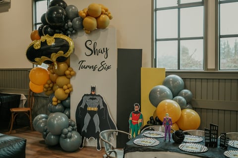 Batman-themed birthday party setup with black and gold balloon arch, superhero decorations, and table settings with comic book characters
