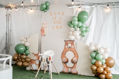 Woodland-themed birthday party setup with Koye is a Big One sign, forest animal cutouts, and green, white, and gold balloon clusters