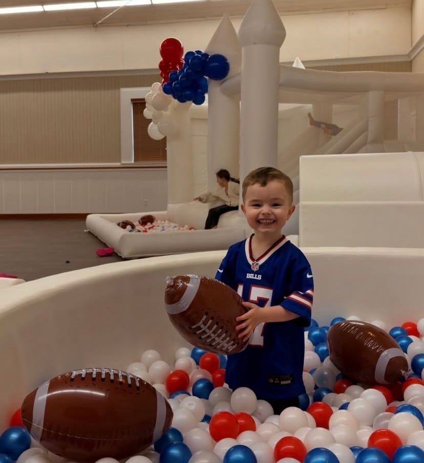 Young child in Bills jersey standing in ball pit filled with red, white, and blue balls holding inflatable football
