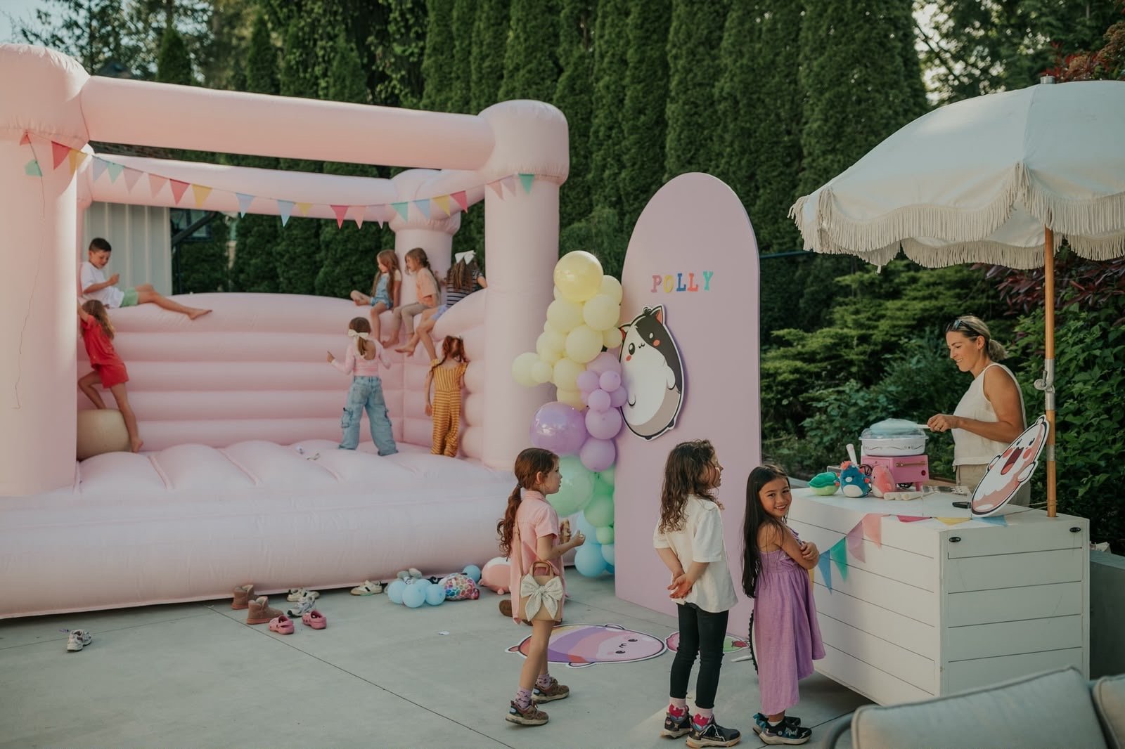 Children playing at an outdoor birthday party with a pink inflatable bounce house, colorful balloons, and a dessert station under an umbrella