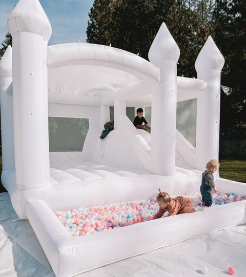 White inflatable bounce house with turrets and a ball pit filled with colorful balls, children playing inside and outside