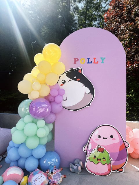 Polly-themed backdrop with rainbow balloons, cute character illustrations, and plush toys in outdoor garden setting