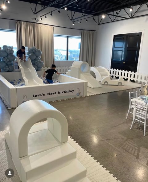Indoor play area with white play structures and obstacles for Levis first birthday party, featuring children playing in a modern venue with windows and industrial ceiling