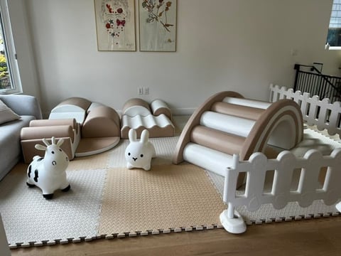Neutral-toned nursery playroom with soft play equipment including a curved climber, white playpen, toy giraffe, bunny, and foam puzzle mat