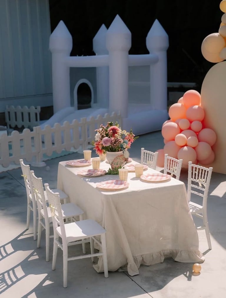 Outdoor childrens party setup with white bounce castle, decorated table with pink settings, white chairs, floral centerpiece, and coral balloon cluster