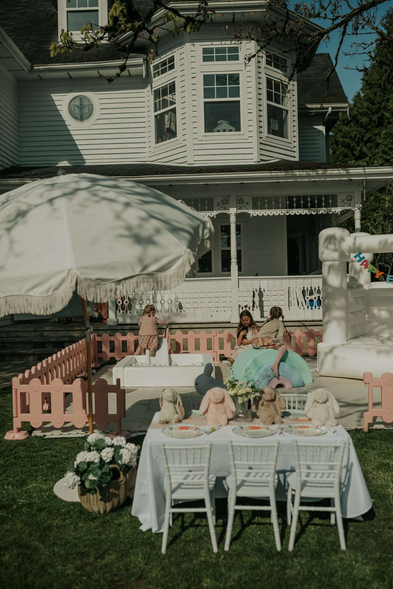 Outdoor doll tea party setup on lawn with miniature furniture, dolls, umbrella, and white Victorian house in background