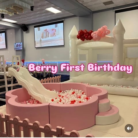 Pink and white inflatable castle bounce house with slide and ball pit labeled Berry First Birthday in an event space