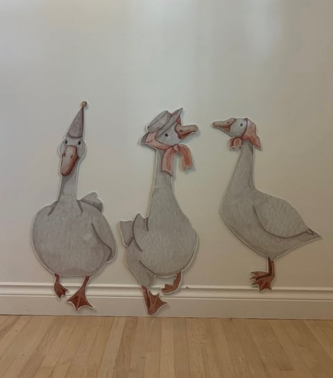 Three whimsical geese illustration in muted gray and red tones displayed on white wall above wooden floor