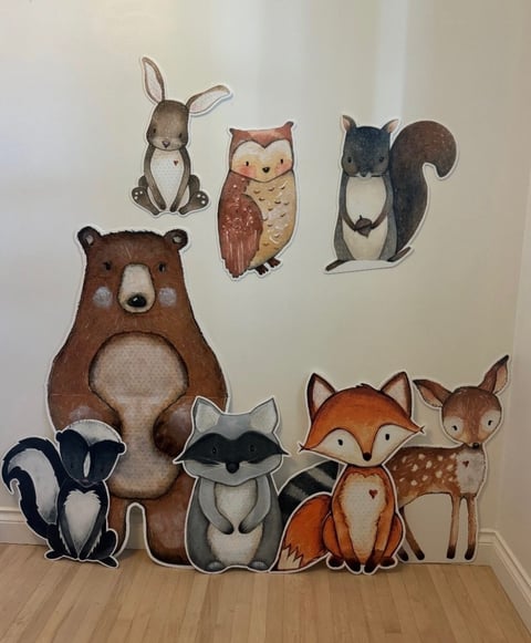 Cardboard cutouts of woodland animals including bear, rabbit, owl, squirrel, skunk, raccoon, fox, and deer arranged on wooden floor