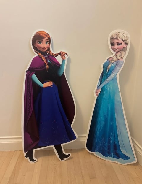 Two Disney Frozen character cardboard cutouts of Anna and Elsa standing on wooden floor against beige wall
