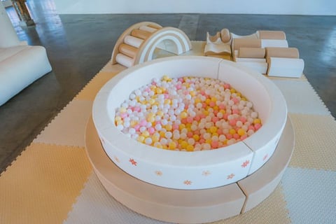 A round white ball pit filled with colorful pink, yellow, and orange plastic balls, surrounded by beige packaging materials and cardboard boxes