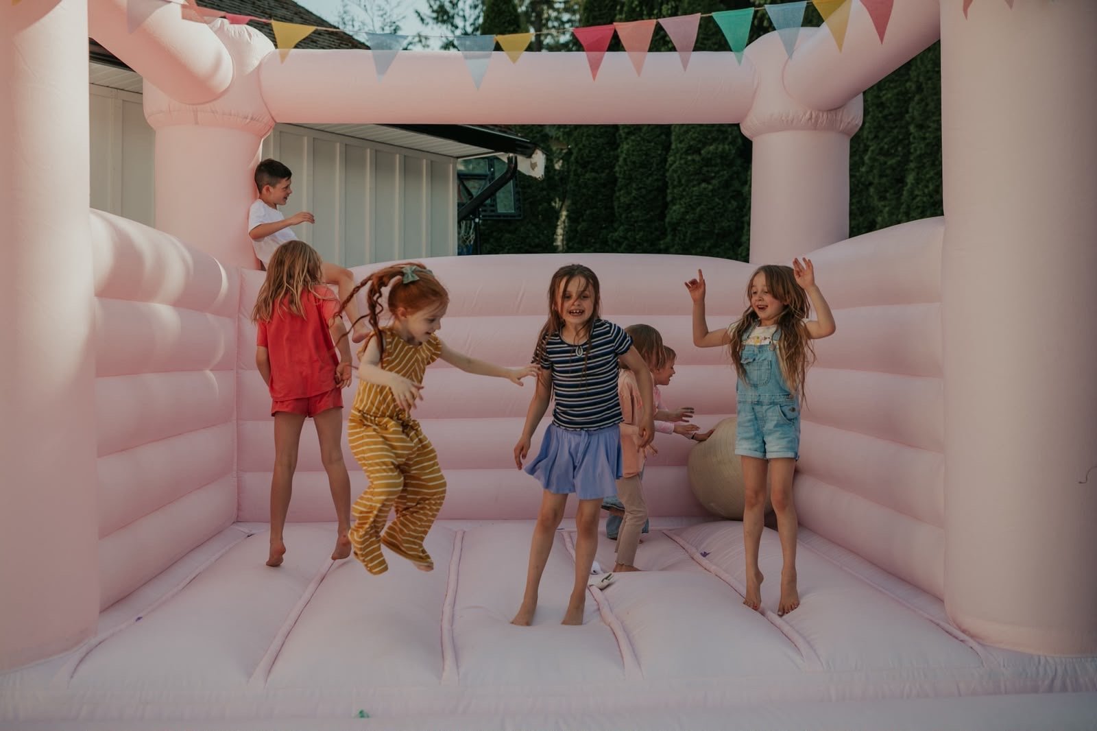 Five children playing and jumping inside a pink inflatable bounce house decorated with colorful bunting and balloons