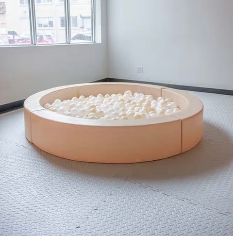Large circular ball pit with peach-colored walls and white rim filled with cream-colored plastic balls in an empty room with windows