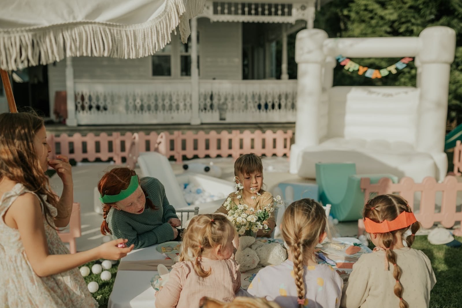 An adult leads outdoor craft activities for a group of children in a backyard with a bounce house and party decorations