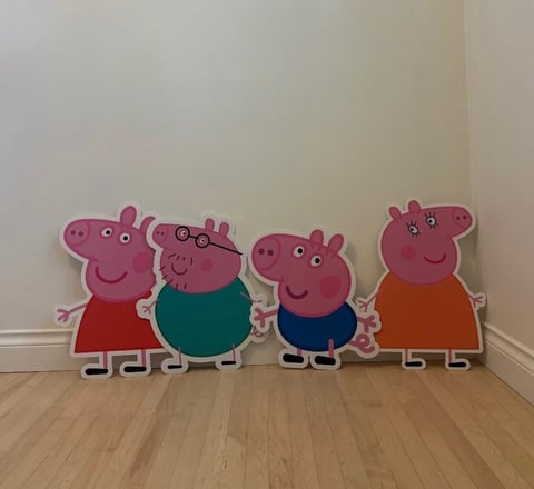 Cardboard cutouts of Peppa Pig characters standing on a wooden floor against a white wall