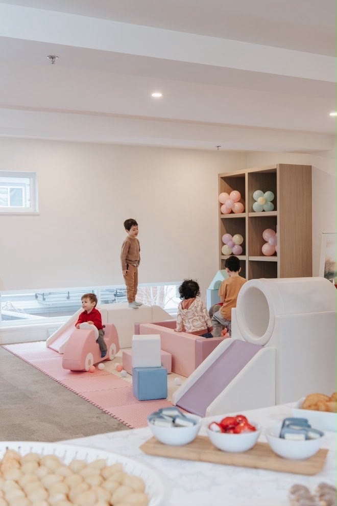 Modern childrens play area with young kids playing with soft pink and blue blocks, shelving unit with colorful balls, and sensory play setup