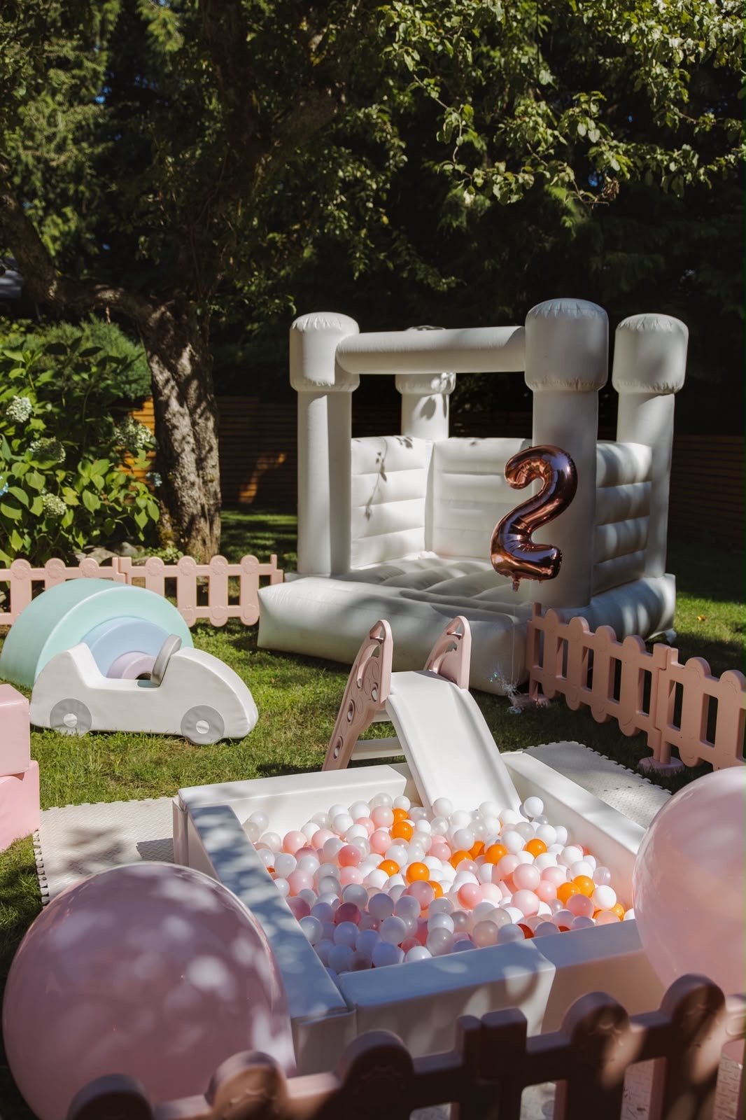 White inflatable bounce house with number 2 in a backyard, surrounded by ball pit, decorative fencing, and pink balloons