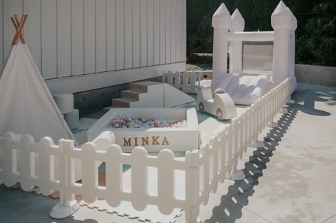 Outdoor childrens play area with white inflatable bounce castle, teepee, ball pit, and MINKA sign surrounded by white picket fence