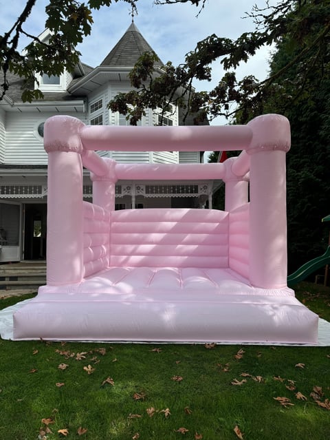 Pink inflatable bounce house with castle towers set up on a lawn in front of a white Victorian house