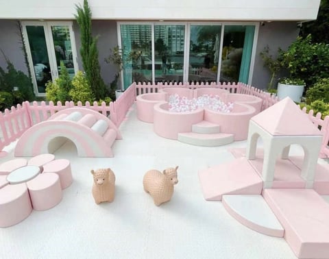 Pink and white playground structure with wooden animal sculptures, enclosed by picket fence, with glass building and greenery in background